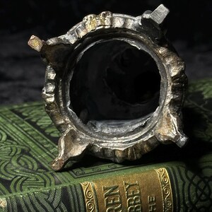 May include: A detailed, antique metal container with a circular opening and ornate, textured detailing. The container has a silver and gold finish, with four small feet. The background features a green book with gold lettering.