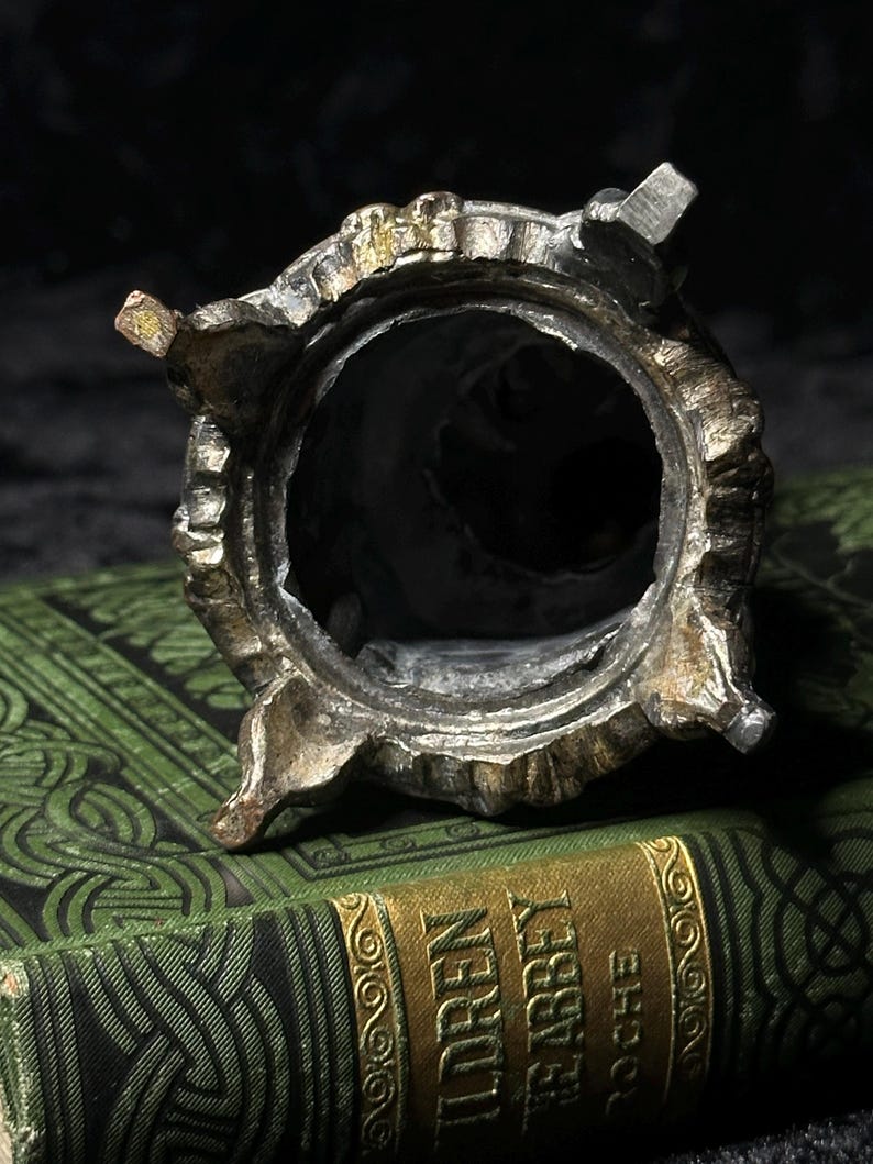 May include: A detailed, antique metal container with a circular opening and ornate, textured detailing. The container has a silver and gold finish, with four small feet. The background features a green book with gold lettering.