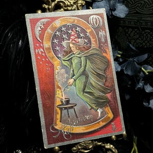 May include: Vintage-style Halloween card featuring a witch in a green dress and cape, wearing a pointed hat. The card has a red and silver border with a crescent moon, stars, and the words "Halloween Ball".