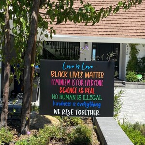 May include: A black sign with colourful text that reads "Love is Love", "Black Lives Matter", "Feminism is for Everyone", "Science is Real", "No Human is Illegal", "Kindness is Everything", and "We Rise Together".