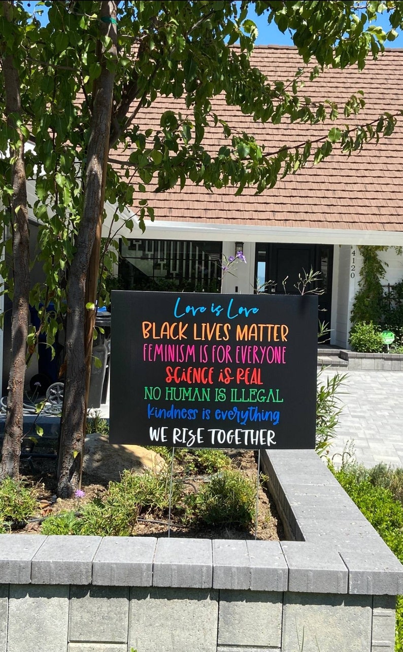 May include: A black sign with colourful text that reads "Love is Love", "Black Lives Matter", "Feminism is for Everyone", "Science is Real", "No Human is Illegal", "Kindness is Everything", and "We Rise Together".