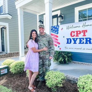 Military Couple in uniform standing in front of personalized “Welcome Home” vinyl banner in front of their home for military celebration.
