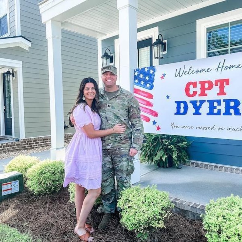 Military Couple in uniform standing in front of personalized “Welcome Home” vinyl banner in front of their home for military celebration.