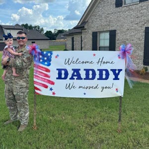 Patriotic “Welcome Home Daddy, We Missed You” banner displayed on lawn for deployment homecoming party. Soldier holding toddler daughter is standing next to the banner