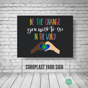 May include: A black and white yard sign with the text "BE THE CHANGE you wish to see IN THE WORLD" in rainbow colors. Two hands, one brown and one white, are forming a heart shape around a globe. The sign also says "COROPLAST YARD SIGN".