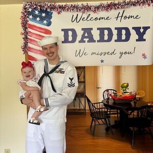 Service member in uniform holding child beside personalized “Welcome Home Daddy, We Missed You” banner for emotional reunion.
