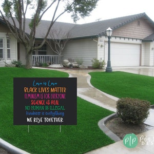 May include: A black yard sign with colourful text that reads "Love is Love, Black Lives Matter, Feminism is for Everyone, Science is Real, No Human is Illegal, Kindness is Everything, We Rise Together".