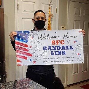 Smiling person displaying personalized “Welcome Home Randal Link” vinyl banner indoors for deployment return. Banner is signed by friends and family with messages