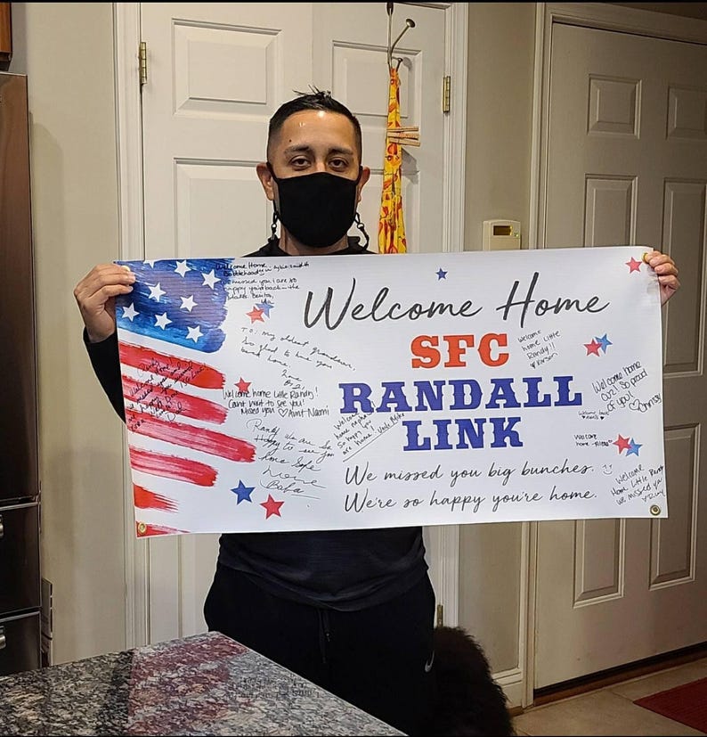 Smiling person displaying personalized “Welcome Home Randal Link” vinyl banner indoors for deployment return. Banner is signed by friends and family with messages