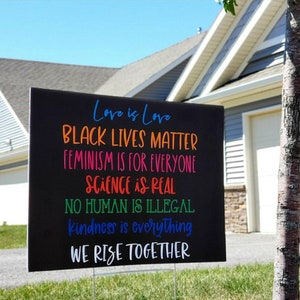 May include: A black sign with colourful text that reads "Love is Love, Black Lives Matter, Feminism is for Everyone, Science is Real, No Human is Illegal, Kindness is Everything, We Rise Together".