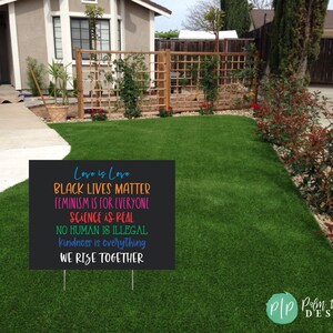 May include: Black yard sign with white text that reads "Love is Love, Black Lives Matter, Feminism is for Everyone, Science is Real, No Human is Illegal, Kindness is Everything, We Rise Together". The sign is on a green lawn.