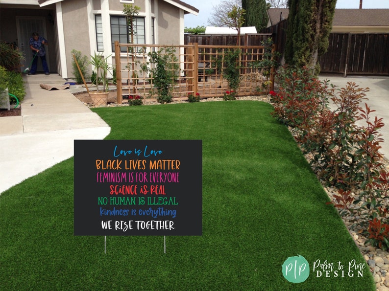 May include: Black yard sign with white text that reads "Love is Love, Black Lives Matter, Feminism is for Everyone, Science is Real, No Human is Illegal, Kindness is Everything, We Rise Together". The sign is on a green lawn.