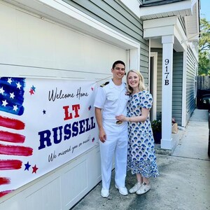 Sailor and Wife standing outside with personalized “Welcome Home” patriotic banner for soldier’s return home hung on garage door. American Flag in the background