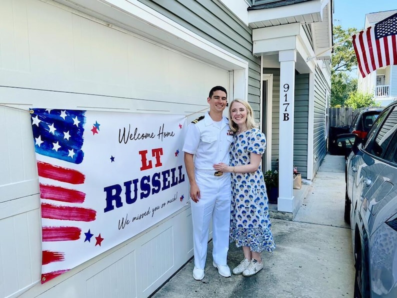 Sailor and Wife standing outside with personalized “Welcome Home” patriotic banner for soldier’s return home hung on garage door. American Flag in the background