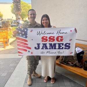 Service member in uniform and partner holding personalized “Welcome Home SSG James” banner for emotional reunion.