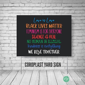 May include: Black yard sign with the text "Love is Love", "Black Lives Matter", "Feminism is for Everyone", "Science is Real", "No Human is Illegal", "Kindness is Everything", and "We Rise Together". The sign is against a white brick wall background.