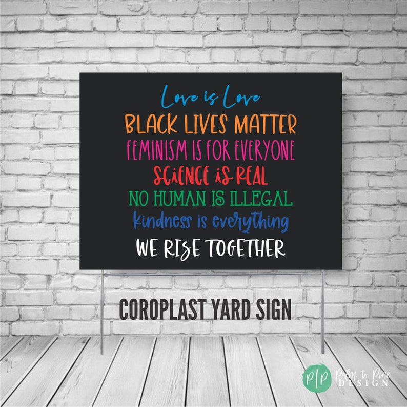 May include: Black yard sign with the text "Love is Love", "Black Lives Matter", "Feminism is for Everyone", "Science is Real", "No Human is Illegal", "Kindness is Everything", and "We Rise Together". The sign is against a white brick wall background.