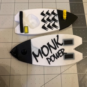 May include: Two white and black surfboard-shaped keychains. One keychain has a black and white design of a person surfing. The other keychain has the words "Monk Power" written in black on a white background.