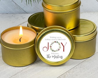 May include: Gold-colored candle tins, some with lids, are arranged on a white wooden surface. One candle is lit, showing a flame. The label on one tin reads "Wishing you a holiday season filled with JOY" with a wreath design and the name "The Martins."