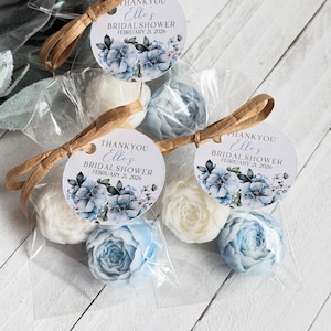 May include: Clear plastic bags containing white and light blue flower-shaped soaps, each tied with a brown raffia ribbon and a round tag reading THANK YOU Elle's BRIDAL SHOWER FEBRUARY 21, 2026, featuring a blue floral watercolor illustration.