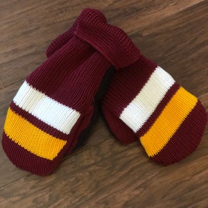 May include: A pair of maroon knitted mittens with white and gold stripes. The mittens are folded together and are lying on a wooden surface.