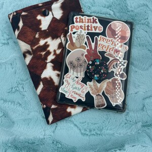May include: A brown and white cowhide-patterned book cover next to a black tablet case. The tablet case is decorated with stickers that say "think positive" and "you're so golden". A black pop socket is attached to the case.