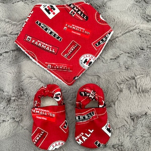 May include: A red baby bib and matching baby shoes. The bib and shoes are patterned with the McCormick Farmall logo and text. The bib is square with white trim. The shoes have a soft sole and elastic ankle strap.