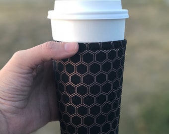 May include: A black and rose gold coffee cup sleeve with a honeycomb pattern. The sleeve is made of neoprene and is designed to keep drinks hot or cold.