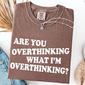 Op de afbeelding: Een bruin t-shirt met de tekst "ARE YOU OVERTHINKING WHAT I'M OVERTHINKING?" wordt getoond met een blauwe jeans, een zilveren ketting en witte instappers. Een gedroogd palmblad is ook zichtbaar.