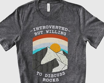 Könnte beinhalten: Ein dunkel-helles graues T-Shirt mit einer Grafik einer Bergkette und einem Sonnenuntergang. Der Text "Introvertiert, aber bereit, über Steine zu diskutieren" ist auf das T-Shirt gedruckt.
