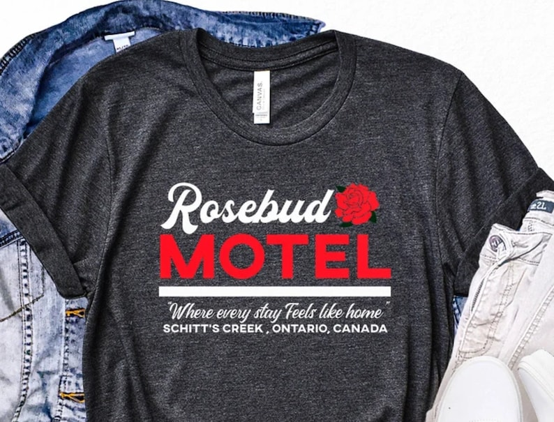 May include: A dark grey t-shirt with a white graphic of the words "Rosebud Motel" in a retro font. The word "Motel" is in red. A red rose is above the word "Rosebud". The text "Where every stay Feels like home" is below the motel name, and the text "Schitt's Creek, Ontario, Canada" is below that.
