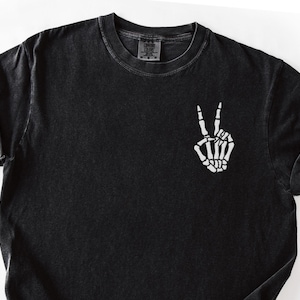 K&ouml;nnte beinhalten: Schwarzes T-Shirt mit einer wei&szlig;en Skeletthand, die ein Friedenszeichen zeigt. Das Shirt hat einen Rundhalsausschnitt und kurze &Auml;rmel mit umgeschlagenen Manschetten. Der Stoff hat einen verblichenen Vintage-Look.