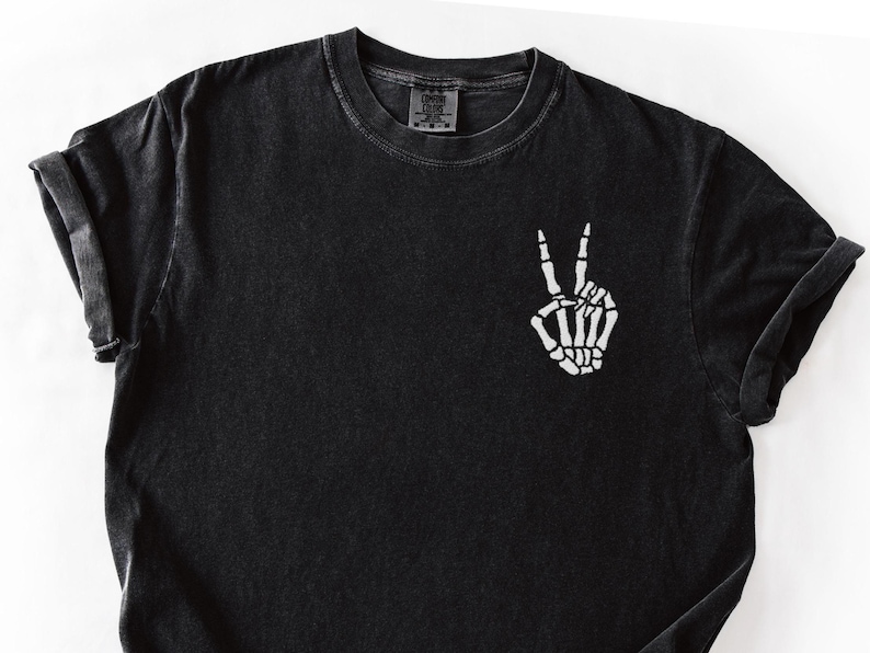 K&ouml;nnte beinhalten: Schwarzes T-Shirt mit einer wei&szlig;en Skeletthand, die ein Friedenszeichen zeigt. Das Shirt hat einen Rundhalsausschnitt und kurze &Auml;rmel mit umgeschlagenen Manschetten. Der Stoff hat einen verblichenen Vintage-Look.
