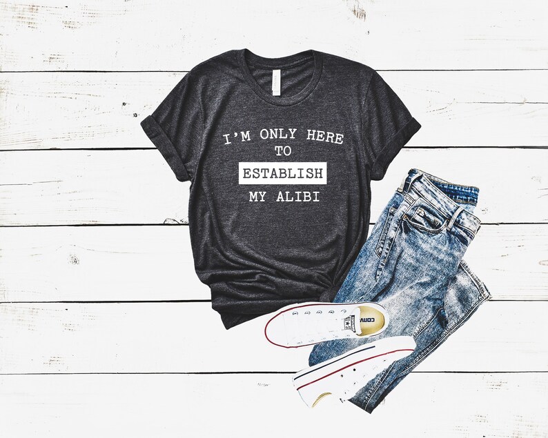 以下が含まれることがあります： 「I'M ONLY HERE TO ESTABLISH MY ALIBI」の白文字がプリントされた濃いグレーのTシャツ。青いジーンズと、赤と黒のアクセントが付いた白いスニーカーが組み合わされています。白い木製の表面に配置されています。