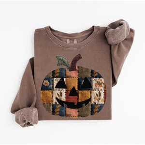 Peut inclure: Sweat-shirt marron avec un motif citrouille en patchwork. La citrouille a un visage souriant et est compos&eacute;e de divers carr&eacute;s de tissu avec des motifs floraux et g&eacute;om&eacute;triques. Les manches ont un revers roul&eacute;.