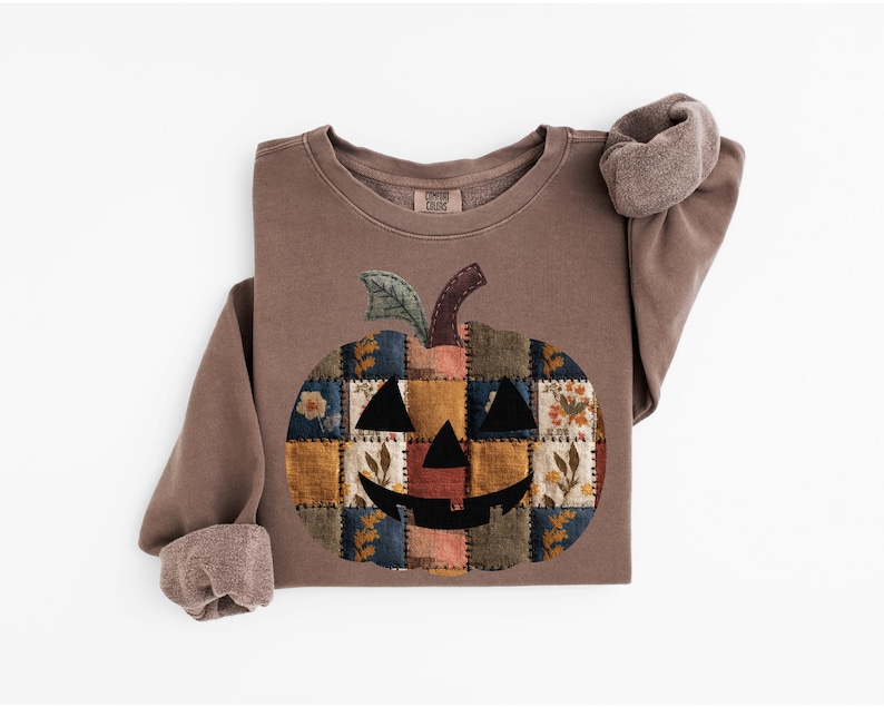 Peut inclure: Sweat-shirt marron avec un motif citrouille en patchwork. La citrouille a un visage souriant et est compos&eacute;e de divers carr&eacute;s de tissu avec des motifs floraux et g&eacute;om&eacute;triques. Les manches ont un revers roul&eacute;.