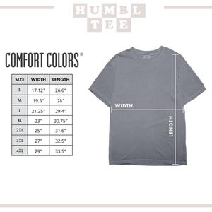 Op de afbeelding: Een grijs Comfort Colors t-shirt met maat- en meetgegevens. De breedte en lengte van het shirt worden met pijlen aangegeven. De maattabel toont de afmetingen in cm.