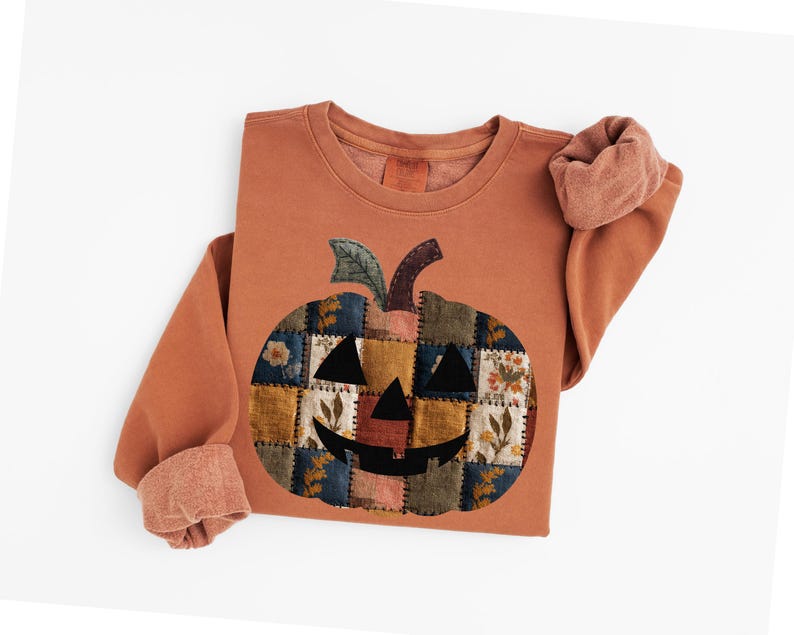 Peut inclure: Sweat-shirt de couleur rouille avec un motif de citrouille en patchwork. La citrouille a un aspect cousu avec divers carr&eacute;s de tissu dans les tons orange, jaune, bleu et vert. Les manches sont retrouss&eacute;es, r&eacute;v&eacute;lant un tissu plus clair et textur&eacute;.