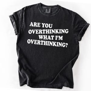 Op de afbeelding: Zwart T-shirt met de witte tekst: "ARE YOU OVERTHINKING WHAT I'M OVERTHINKING?" Het T-shirt heeft een ronde hals en opgerolde mouwen. De stof heeft een vervaagde, vintage uitstraling. Een casual kledingstuk.