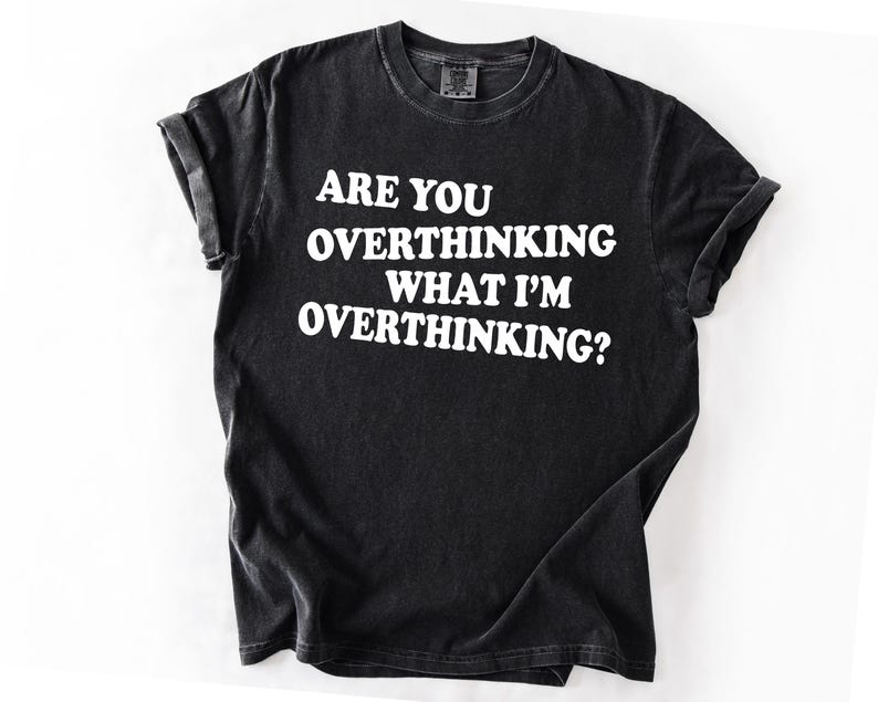 Op de afbeelding: Zwart T-shirt met de witte tekst: "ARE YOU OVERTHINKING WHAT I'M OVERTHINKING?" Het T-shirt heeft een ronde hals en opgerolde mouwen. De stof heeft een vervaagde, vintage uitstraling. Een casual kledingstuk.