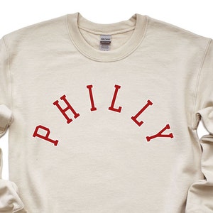 May include: A cream-colored sweatshirt with a red "PHILLY" graphic printed on the front.