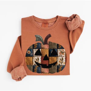 Peut inclure: Sweat-shirt de couleur rouille avec un motif de citrouille en patchwork. La citrouille a un visage souriant et est compos&eacute;e de divers carr&eacute;s de tissu avec des motifs floraux et g&eacute;om&eacute;triques. Les manches sont retrouss&eacute;es.