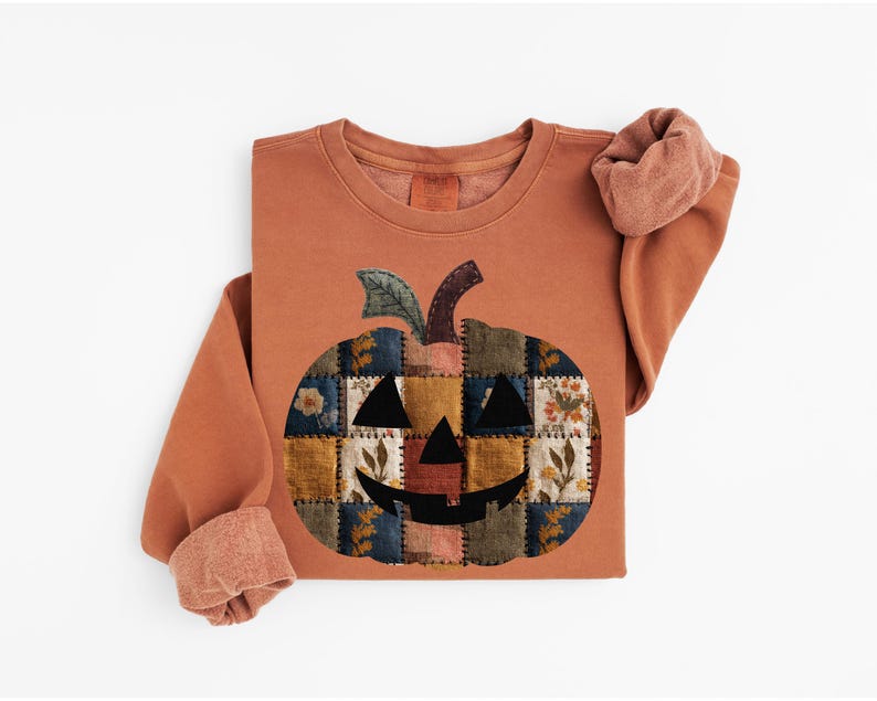 Peut inclure: Sweat-shirt de couleur rouille avec un motif de citrouille en patchwork. La citrouille a un visage souriant et est compos&eacute;e de divers carr&eacute;s de tissu avec des motifs floraux et g&eacute;om&eacute;triques. Les manches sont retrouss&eacute;es.