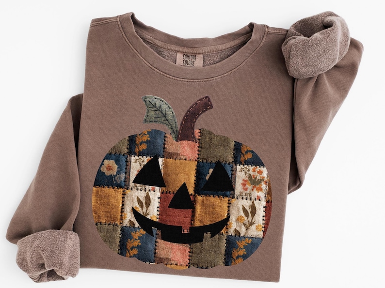 Peut inclure: Un sweat-shirt marron avec un motif de citrouille en patchwork. La citrouille est compos&eacute;e de divers carr&eacute;s de tissu aux tons orange, bleu et &agrave; motifs floraux. Les manches ont une doublure douce et textur&eacute;e.