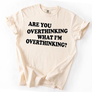 Op de afbeelding: Cr&egrave;mekleurig T-shirt met de tekst "ARE YOU OVERTHINKING WHAT I'M OVERTHINKING?" in vette zwarte letters. Het shirt heeft opgerolde mouwen en een klassieke ronde hals. De stof ziet er zacht en comfortabel uit.