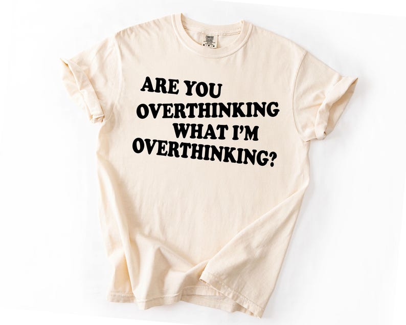 Op de afbeelding: Cr&egrave;mekleurig T-shirt met de tekst "ARE YOU OVERTHINKING WHAT I'M OVERTHINKING?" in vette zwarte letters. Het shirt heeft opgerolde mouwen en een klassieke ronde hals. De stof ziet er zacht en comfortabel uit.