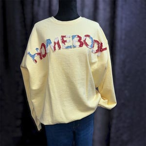 May include: A pale yellow long-sleeve sweatshirt featuring the word HOMEBODY appliqued across the chest in a textured, frayed fabric with a red, white, and blue floral pattern. The garment has a crew neckline and a relaxed, loose-fitting silhouette.