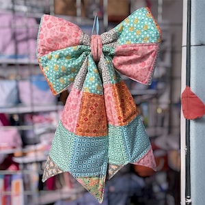 May include: A large, colorful quilted bow with a patchwork design. The bow features various patterned fabrics in shades of pink, teal, orange, and green. It is suspended by a light blue ribbon, showcasing a handmade, decorative item.