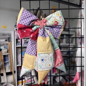May include: A large, handmade quilted bow in a variety of fabrics and patterns. The bow features a patchwork design with floral, geometric, and gingham prints in shades of pink, purple, yellow, and green. The bow is hanging from a black metal grid.