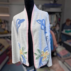 May include: A white open-front cardigan with a textured wavy pattern, featuring light pink trim along the edges. The garment is decorated with embroidered floral motifs in shades of light blue, soft yellow, and sage green on the front panels.